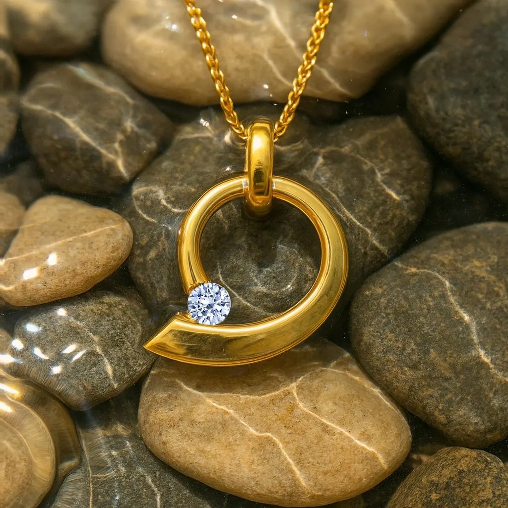 A minimalist yellow gold circular pendant featuring a brilliant-cut white diamond, photographed underwater resting on smooth river pebbles.