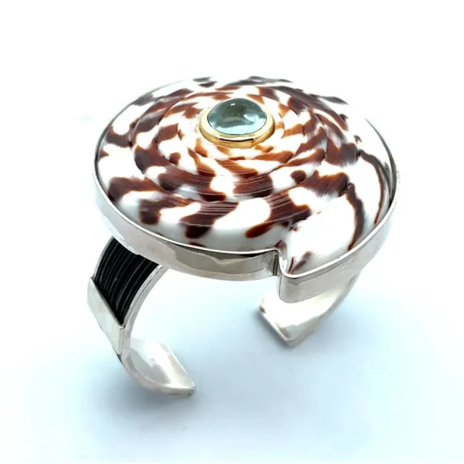 Artisan silver cuff bracelet featuring a brown and white spiral shell centerpiece with a round blue gemstone inlay.